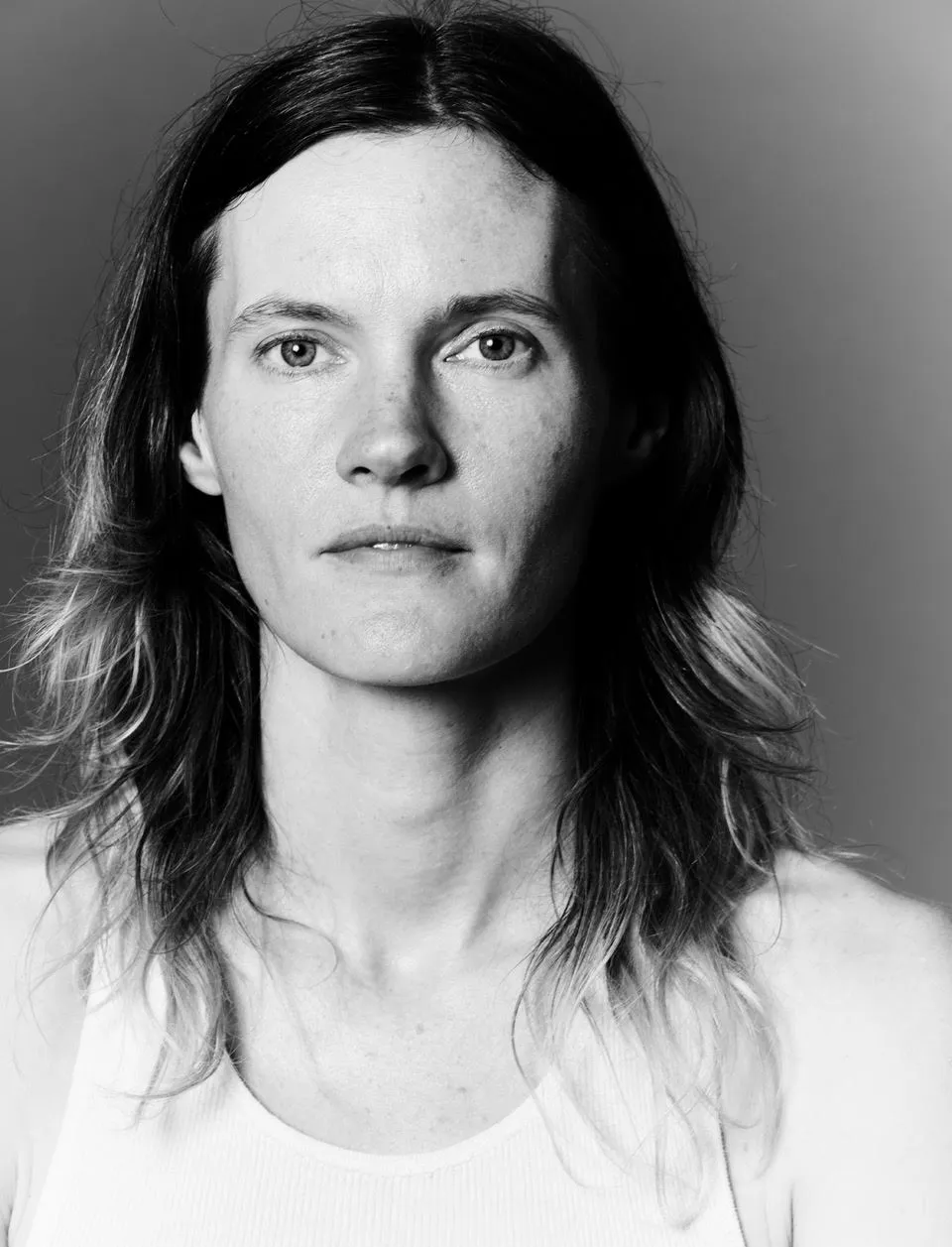 A black and white portrait of a white person with long brown hair wearing a white tshirt